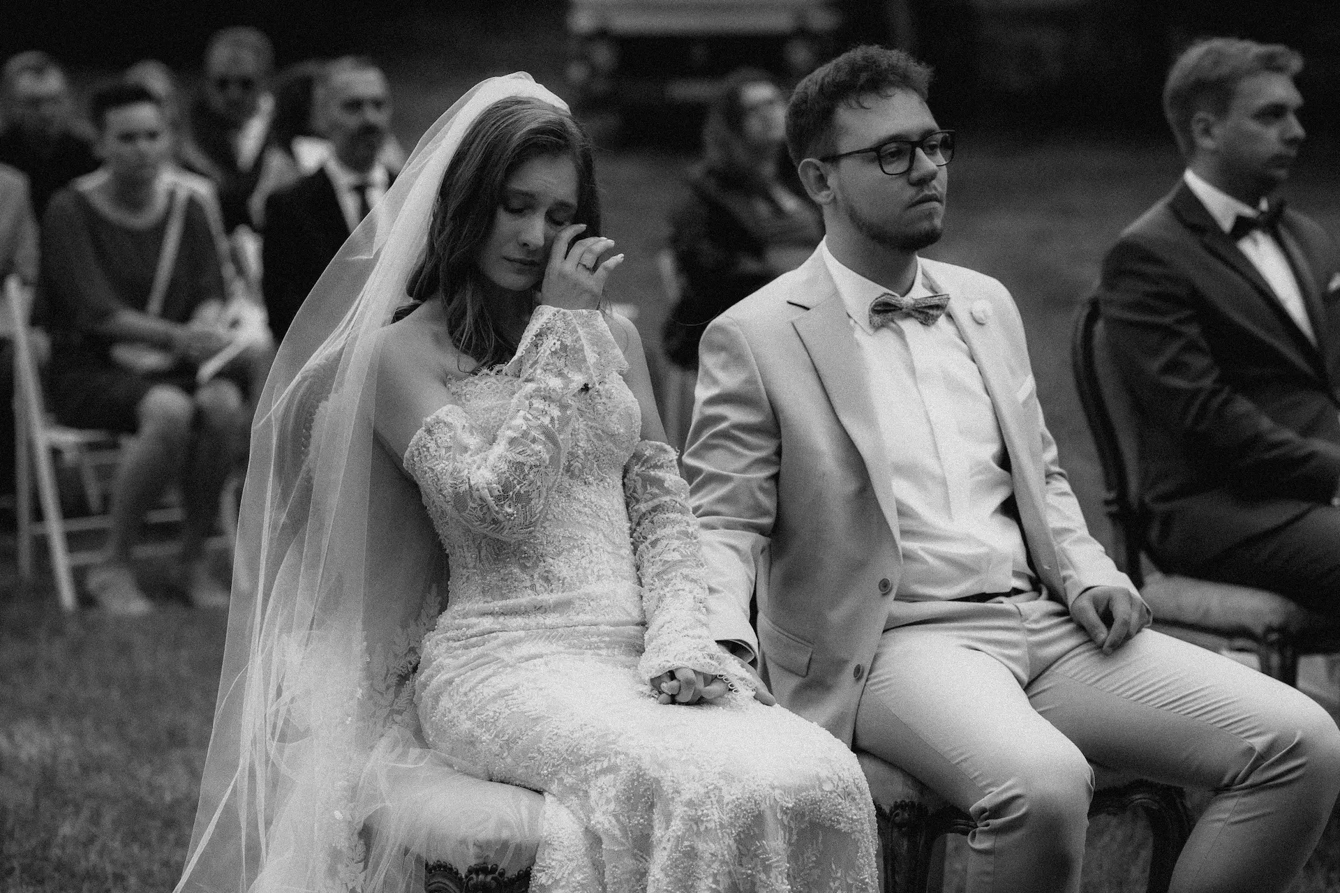 Panna młoda w koronkowej sukni ociera łzy, siedząc obok pana młodego podczas ceremonii plenerowej