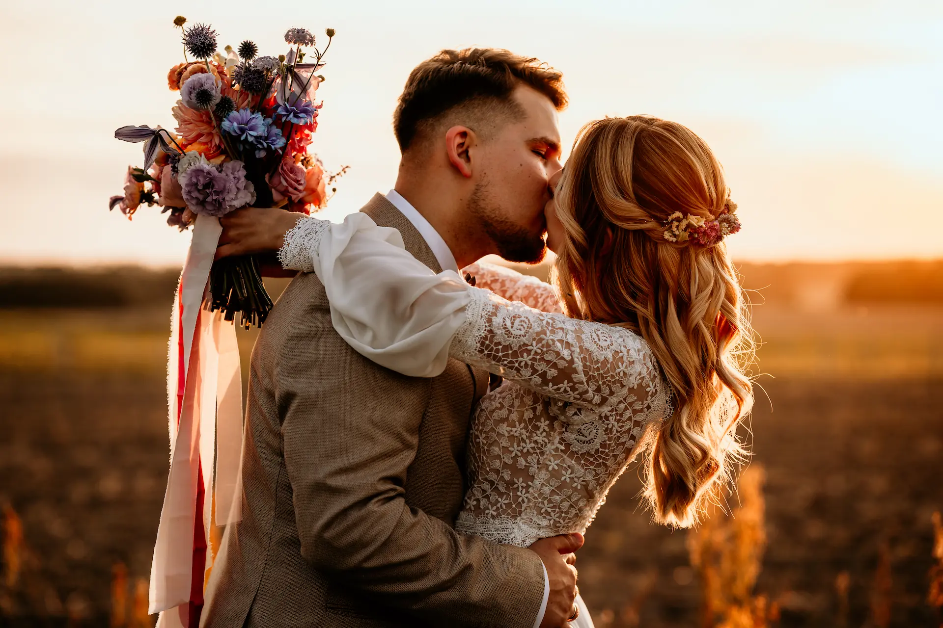 Pan młody całuje pannę młodą trzymającą kolorowy bukiet w promieniach zachodu słońca.